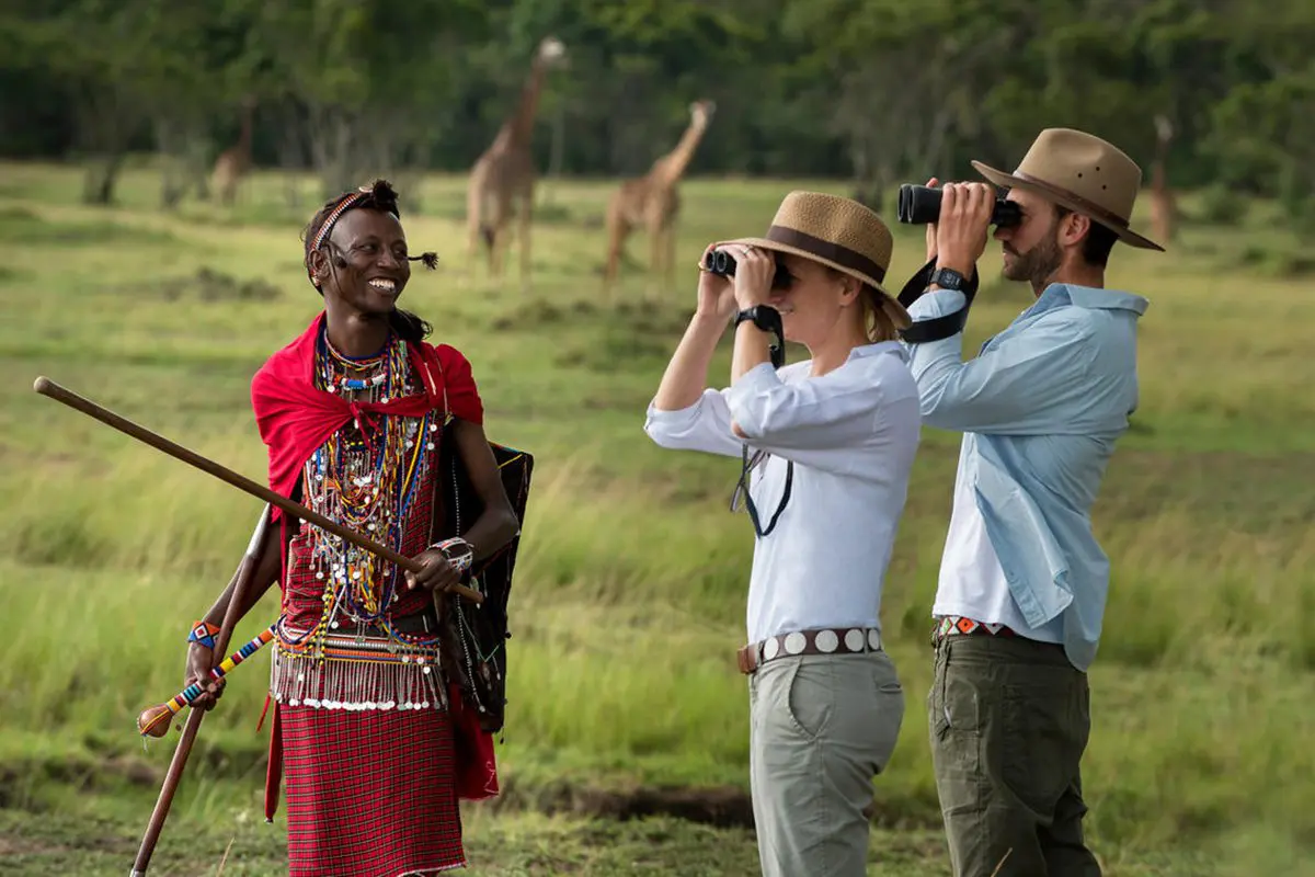 Two travellers holding up binoculars and their guide out on a walking safari with two giraffe in the background | Go2Africa