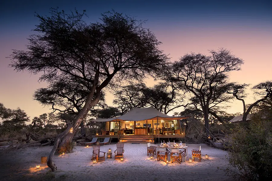 Una estructura principal de carpas rodeada de árboles se asoma a las mesas y sillas preparadas para comer, con farolillos que iluminan la oscuridad casi total | Go2Africa