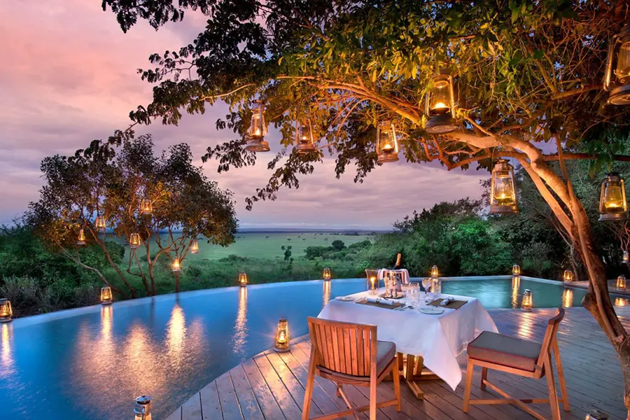 A table for two set up under a tree by a curved infinity pool while the tree above and adjacent, as well as the pool, are adorned with lanterns | Go2Africa