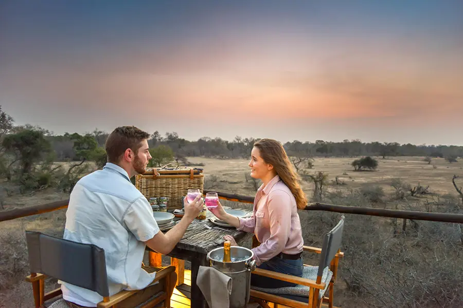 Una pareja sentada en una mesita chocando sus copas mientras contemplan una vista panorámica de Kruger | Go2Africa