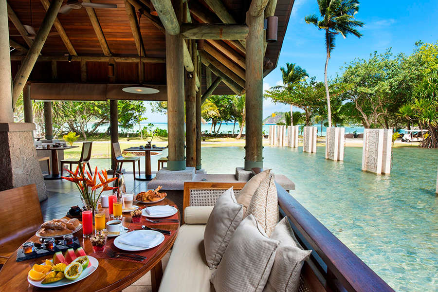 A breakfast table to the left on an elevated wood structure surrounded by a body of water with a beach and the ocean beyond | Go2Africa