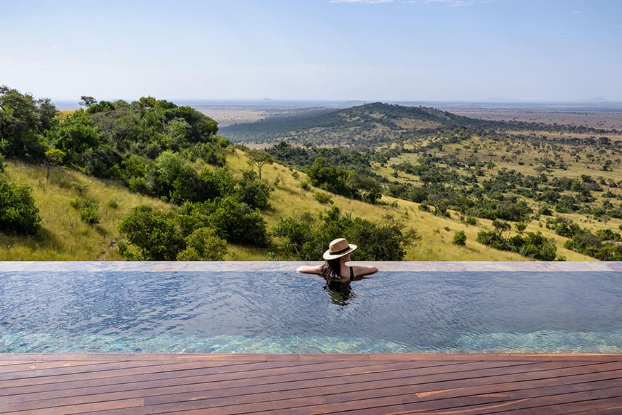 Una mujer mira de espaldas a la cámara con sombrero mientras se sumerge hasta los hombros en una piscina con vistas a un valle | Go2Africa
