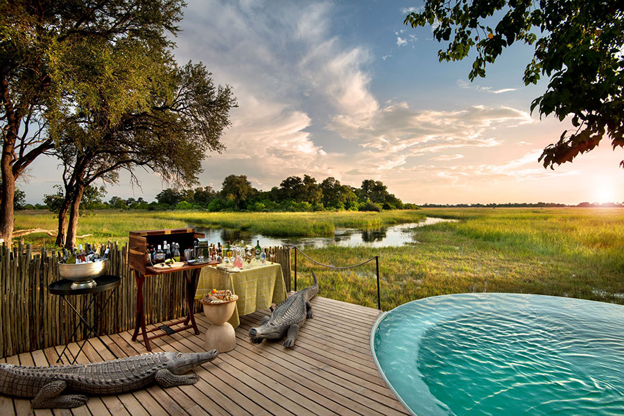 Part of a pool, two fake crocodiles and a portable bar on a deck overlooking a river and lush grass and trees | Go2Africa