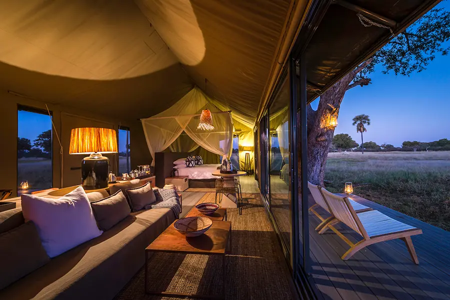 Vista de una suite en una tienda de campaña, desde el sofá y la mesa de café con una gran lámpara detrás hasta una cama con dosel y mosquitera, mostrando también una pequeña terraza con sillas | Go2Africa