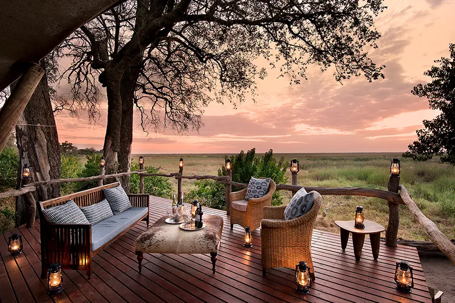 Wicker couch and chairs with a small table in between sits on a wooden deck overlooking vast green plains with a pink sky in the background | Go2Africa