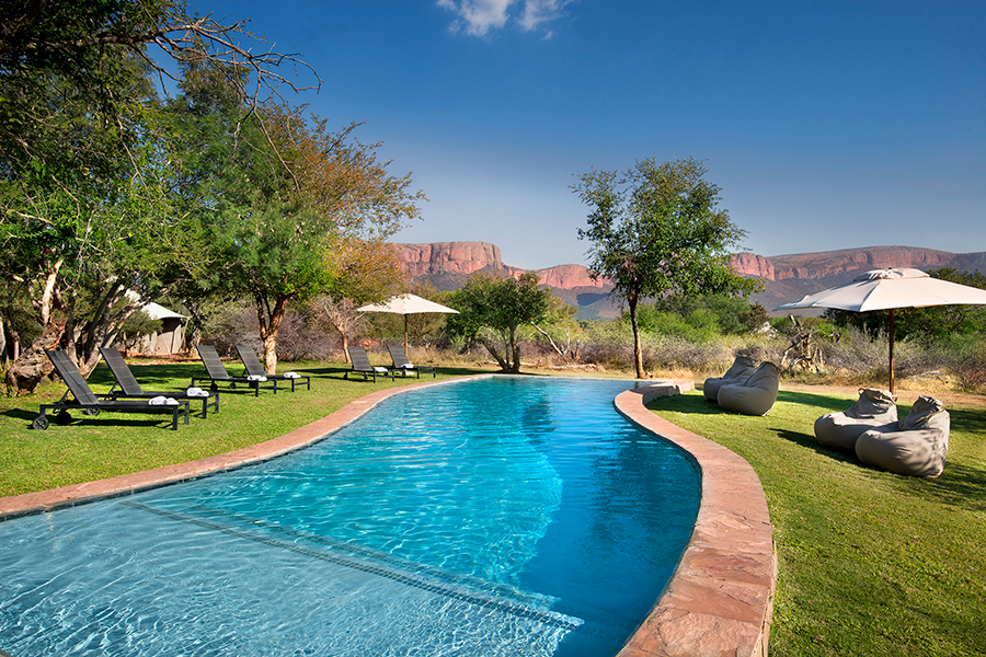 A long sweeping pool surrounded by loungers, umbrellas and trees looks across to a mountain | Go2Africa