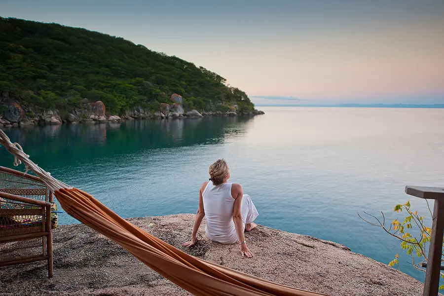 A lady facing away from the camera looks out over Lake Malawi at dusk with an unoccupied hammock behind her | Go2Africa