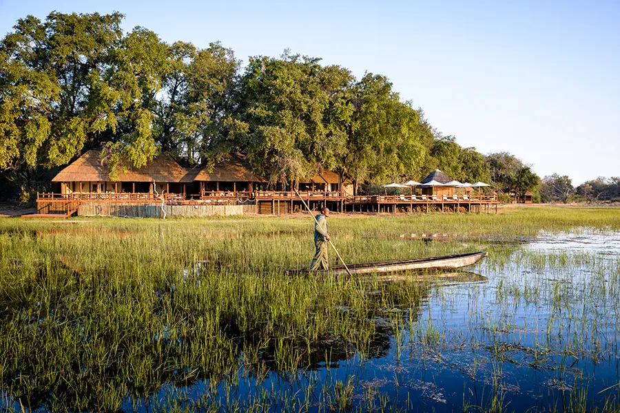 A single mokoro with standing guide moves from right to left along a body of water with a large wooden camp behind it | Go2Africa