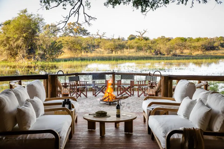 Varios sillones de felpa comparten una mesa de café en la parte delantera de la imagen, mientras que más atrás hay una pequeña hoguera rodeada de sillas de camping con una vista del agua más allá | Go2Africa