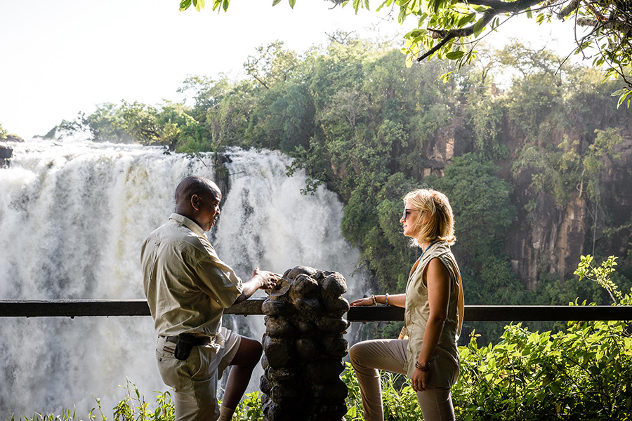 Two people are talking while leaning on a railing overlooking part of Victoria Falls and the surrounding greenery | Go2Africa