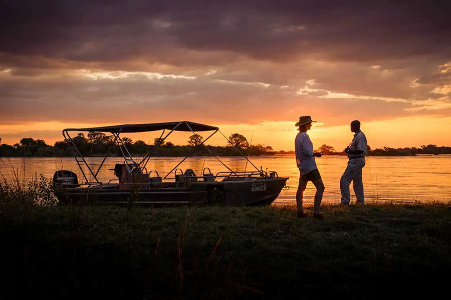 Dos personas de pie en la orilla con un barco con vistas a la puesta de sol | Go2Africa