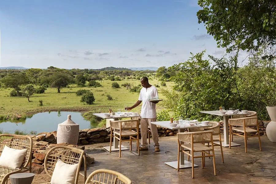 Sillas de mimbre y pequeñas mesas en una terraza elevada con vistas a una masa de agua y a parte de la Reserva de Caza Grumeti | Go2Africa