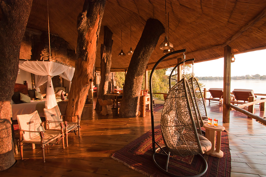 A suite at Tongabezi Lodge in Zambia with matching hanging egg chairs, a large bed, and loungers on the exposed balcony overlooking a body of water | Go2Africa