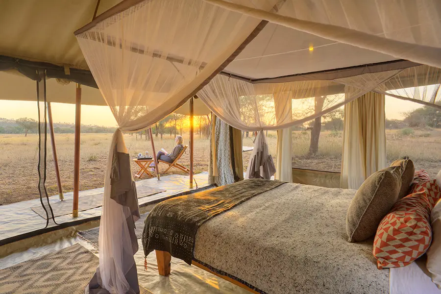 Una mujer sentada en una pequeña mesa y sillas en el patio de una suite en tienda de campaña con una gran cama y mosquitera reunidas en cada esquina | Go2Africa