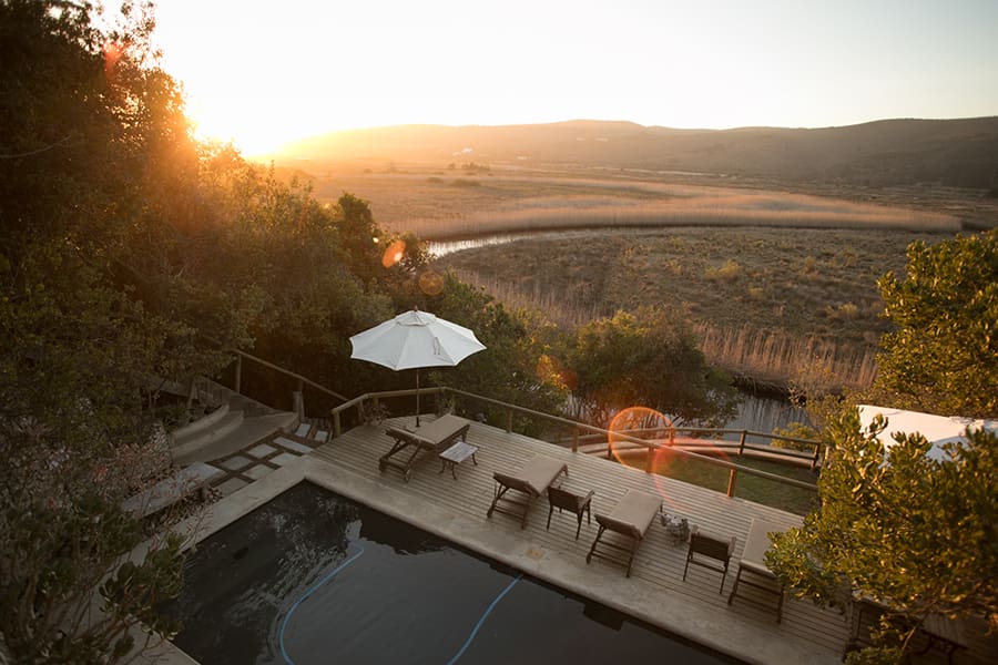 An aerial view over a pool with deck and loungers and down onto the land below, stretching across to the river and sunset in the distance | Go2Africa