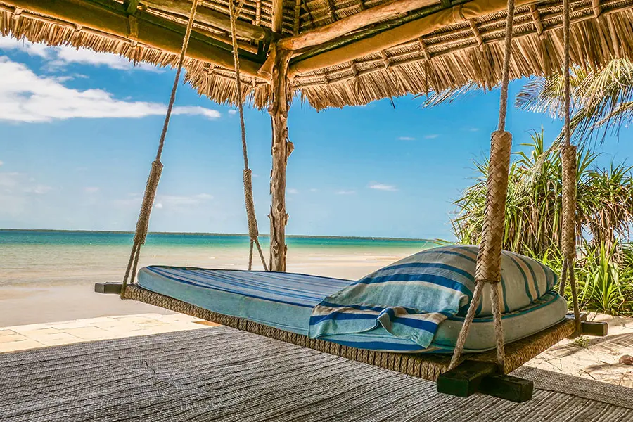 An elevated swinging bed with a pillow and thin mattress sit under a thatched structure on the beach looking at the sea | Go2Africa