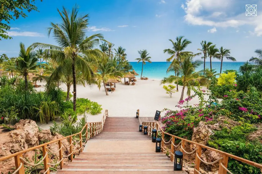 A set of wooden stairs lead down to a white sandy beach complete with palm trees and thatched palapas with the bright blue ocean beyond | Go2Africa