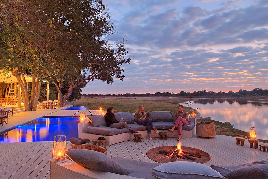 Three guests sit around a fire in a circular boma overlooking a body of water with two pools behind them at Time + Tide Chinzombo