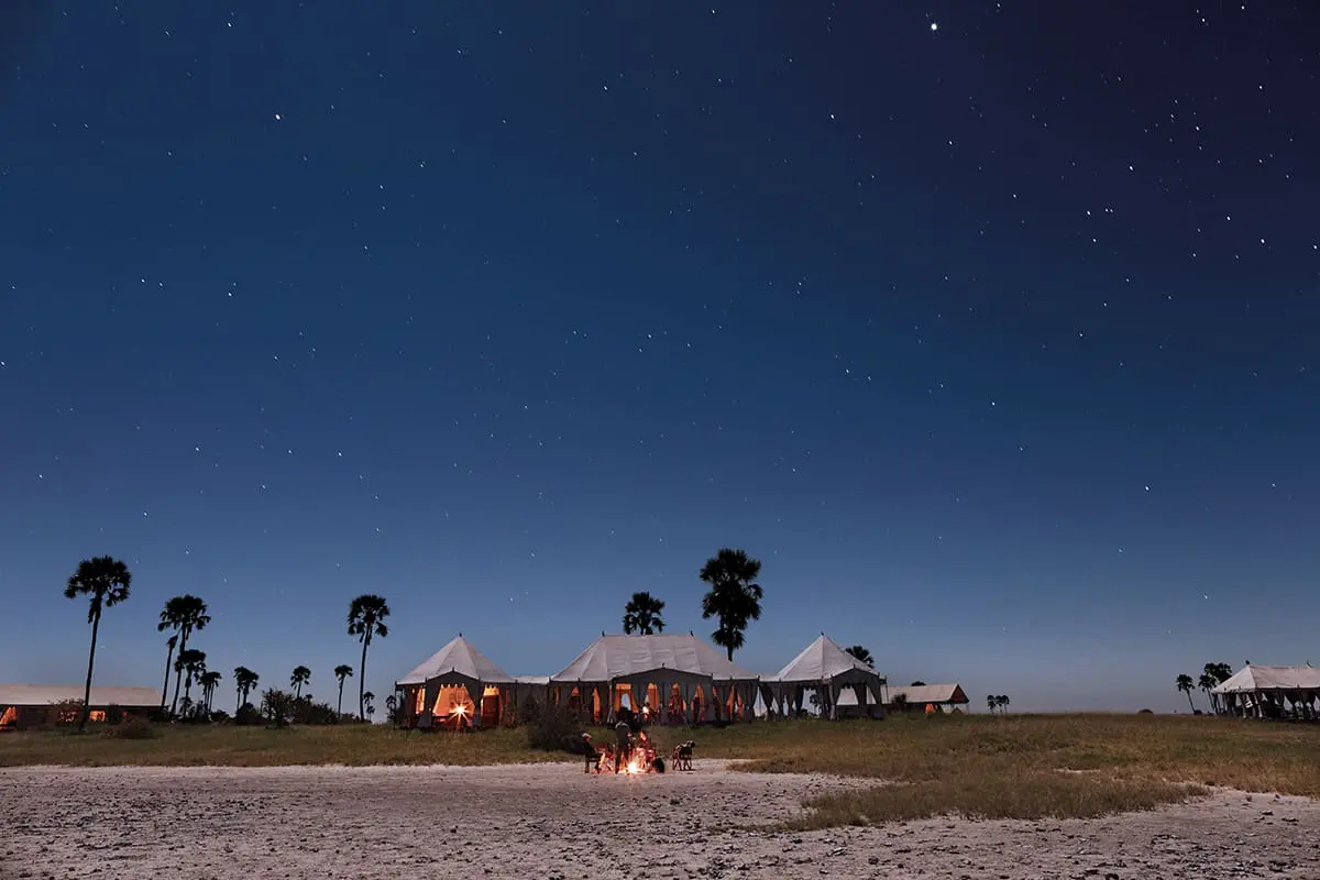 Grupo sentado junto al fuego fuera del Campamento San bajo el cielo nocturno del desierto de Kalahari | Botswana Safari | Go2Africa