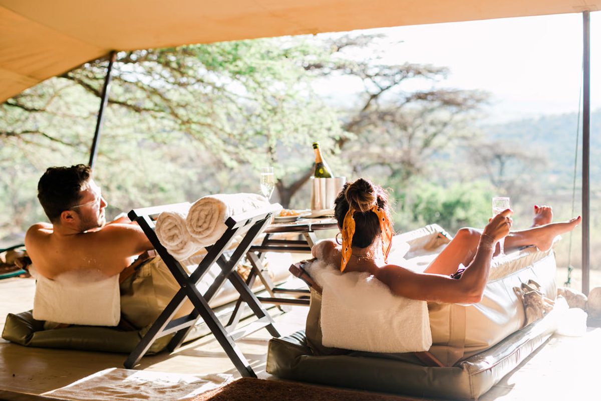 Two people sit in bathtubs underneath a canvas with champagne on the table between them overlooking a lovely view of the bush | Go2Africa