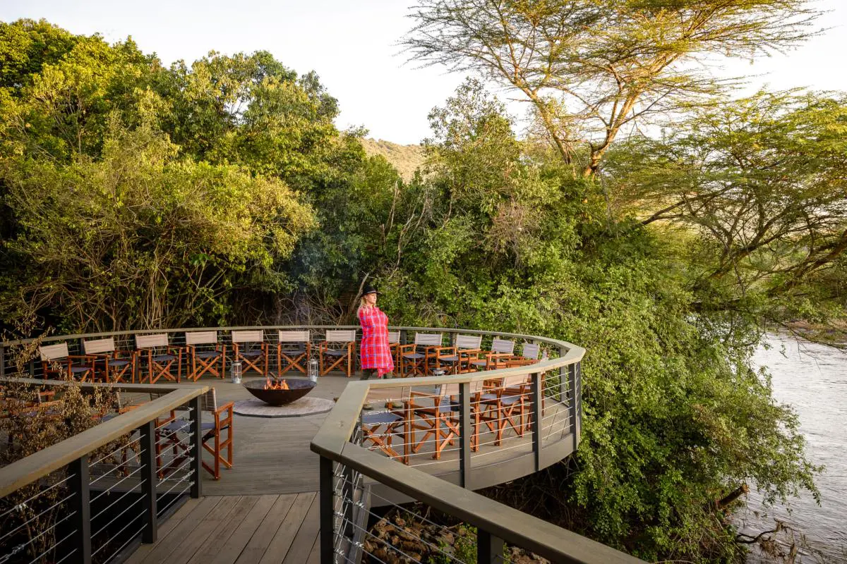 A woman stands in an elevated wooden boma surrounded by camping chairs with an open firepit behind her overlooking the river | Go2Africa