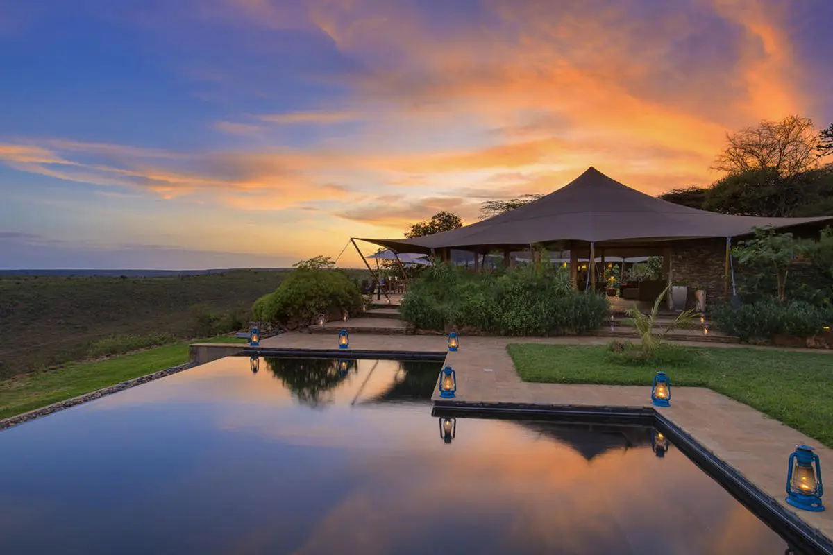 An infinity pool with lanterns around its edges reflect a vivid blue sky with pink and orange clouds across the horizon while a tented structure sits behind the pool | Go2Africa