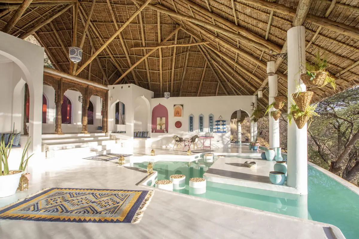 White, marble-like floors contrast with wooden beams supporting a thatched roof above a crystal clear pool with island and pillars | Go2Africa