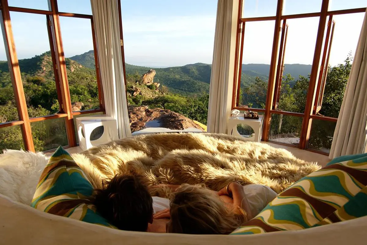 Two people lie under a fluffy rug on a circular bed looking through a wall of windows and out to rolling hills | Go2Africa