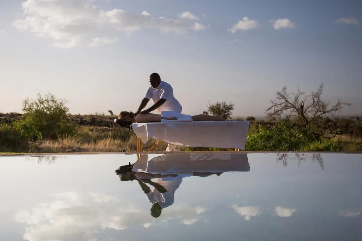 A pristine pool reflects someone receive a massage on a treatment table and the spattering of clouds in the sky, but not the greenery found behind the people | Go2Africa