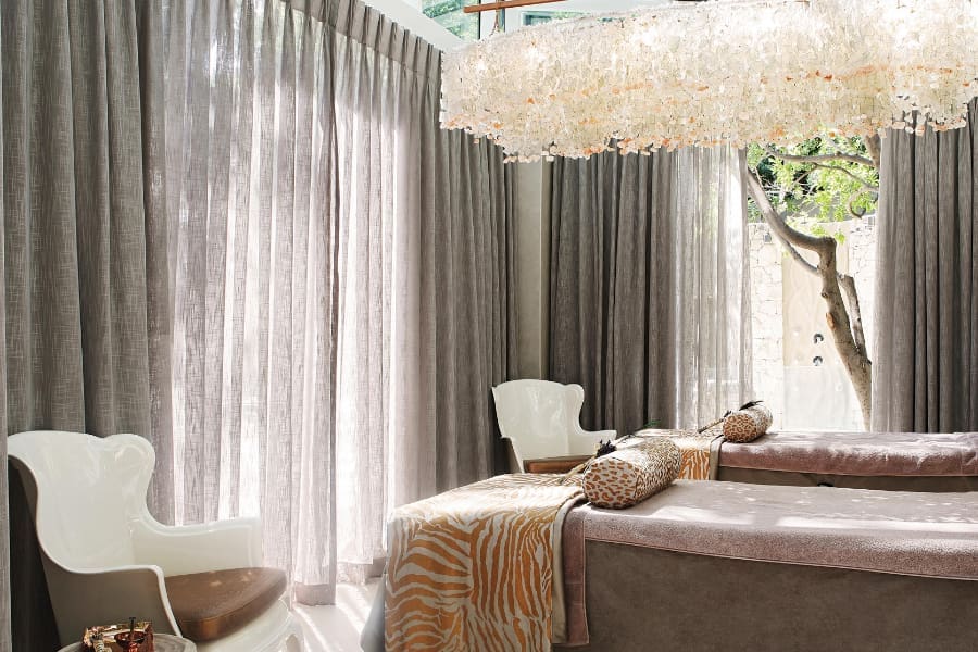 Una sala de tratamiento doble en The Saxon con una hermosa lámpara de araña, cortinas transparentes de color gris claro y un árbol en el exterior | Go2Africa