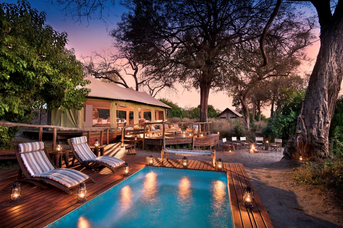 Una terraza elevada con linternas que rodean el agua y una hoguera con sillas de camping y el alojamiento principal en Linyanti Ebony | Go2Africa