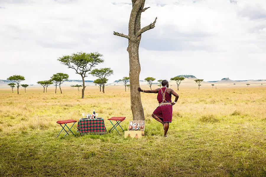 Un guía maasai de pie sobre una pierna mirando a la cámara apoyado en un árbol junto a una cesta de picnic y pequeñas mesas de servicio con escasos árboles de fondo | Go2Africa