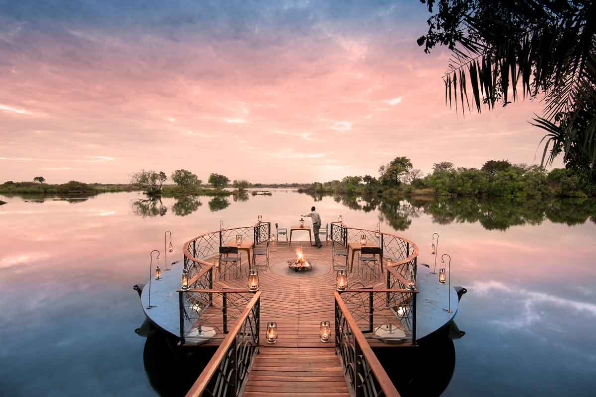 A boma extending a lake features several tables and an open fire pit in the middle with lanterns dotted around the edge lit up by a dusky sky, which is perfectly reflected in the surrounding water | Go2Africa