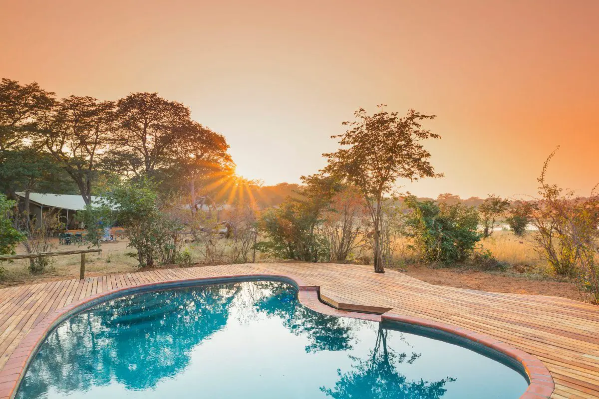 Una piscina en forma de riñón rodeada de terrazas de madera con vistas a los arbustos de los alrededores y a una puesta de sol en la distancia en Verney's Camp en Hwange | Go2Africa