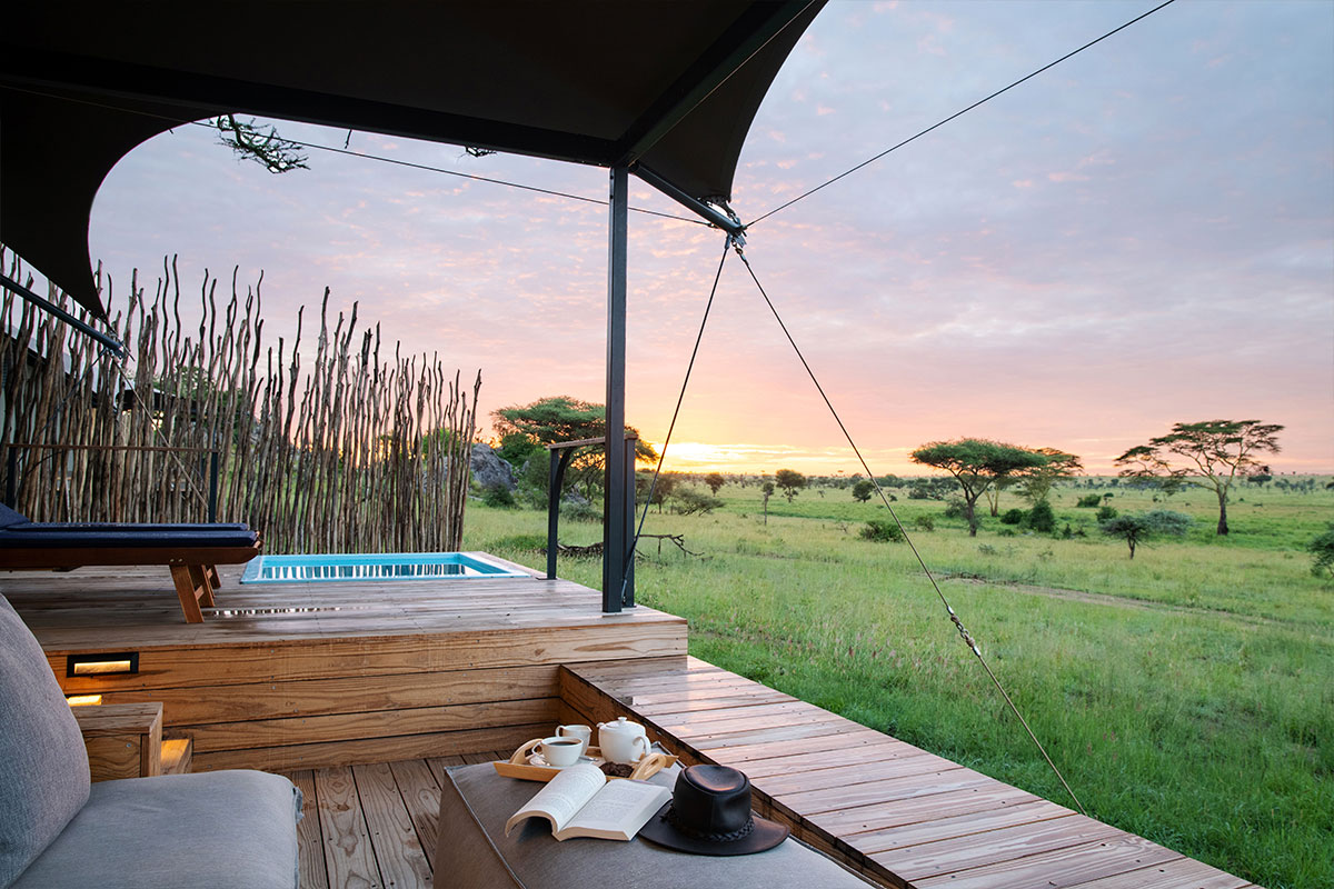 A wooden deck with a raised plunge pool and indented seating section featuring a coffee table with book, hat, and refreshment tray overlook vast grasslands and across to a sunlit skyline | Go2Africa