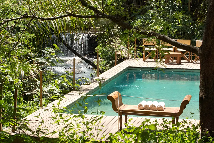 A look through trees and shrubbery across to a pristine pool on a raised deck with a small seating area that all overlooks an adjacent river found a level below | Go2Africa