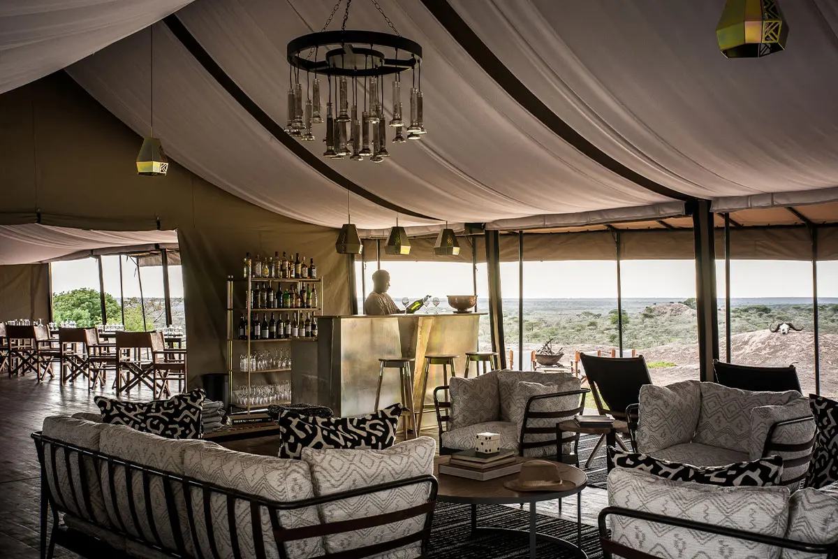 Una sala de estar con bar y lámpara de araña con vistas al Serengeti desde el campamento Sanctuary Kichakani | Go2Africa