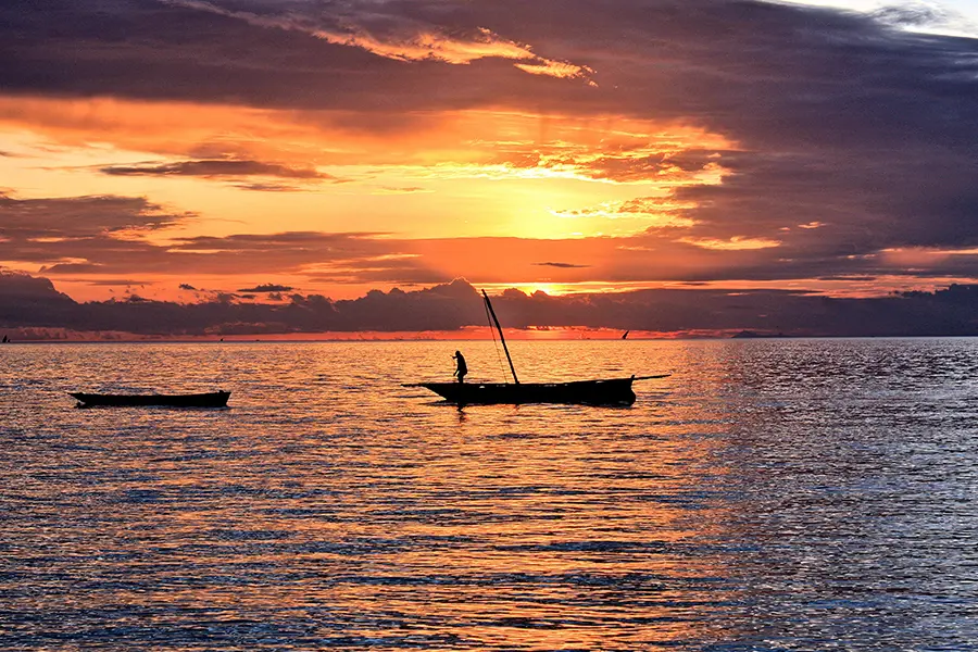 A dhow sails on the waters of Zanzibar at sunset, and orange sky creating glimmers of warm light on the water's surface