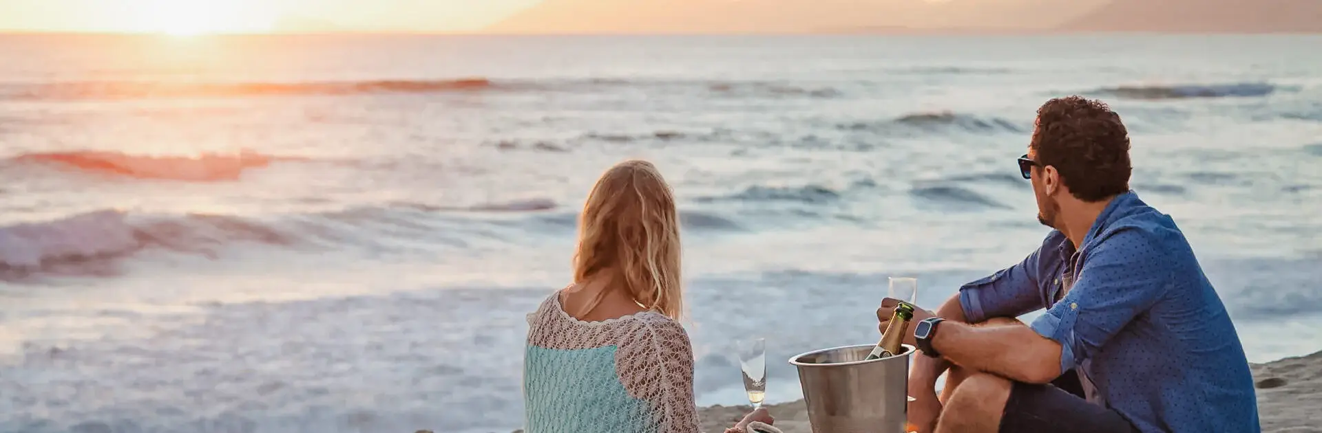 Una pareja sentada en la playa mirando al océano con copas de champán y una cubitera con la botella entre los dos | Go2Africa