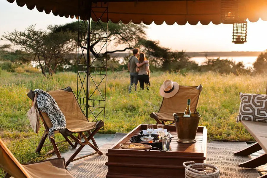 Un hombre y una mujer observan a lo lejos la puesta de sol en un safari mientras en primer plano hay una zona al aire libre con vino y aperitivos | Go2Africa