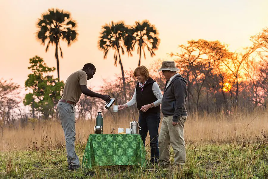 Two guests stand to the right of a small serving table while their guide pours the contents of a jug into one of their cups while the sun sets behind them | Go2Africa