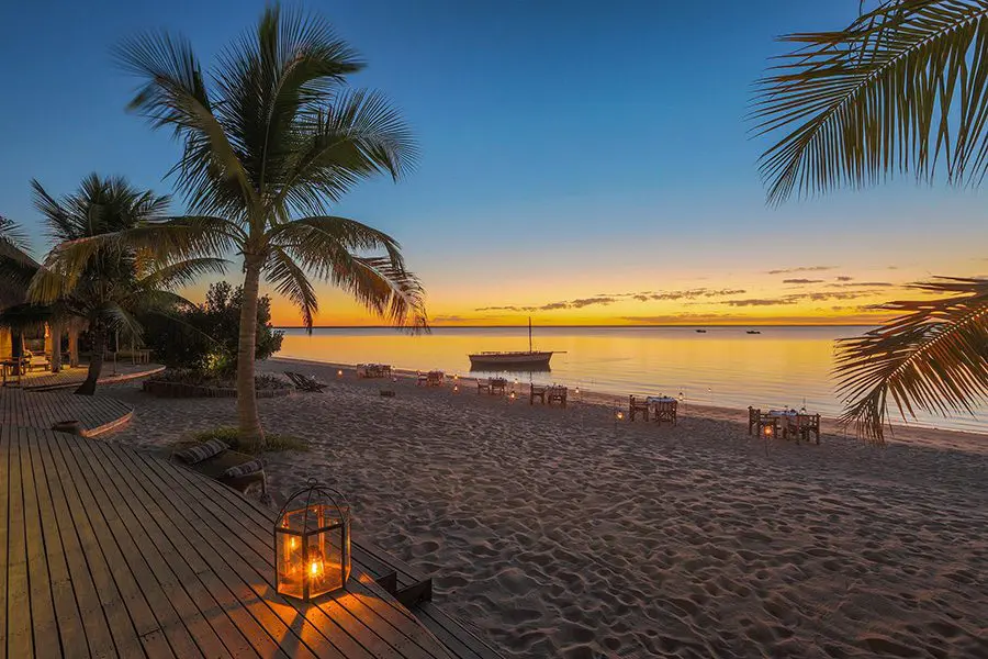 Several small lantern lit tables sit on the beach near the water with palm trees either side overlooking the last of an orange sunset with a small boat docked in the shallow waters | Go2Africa