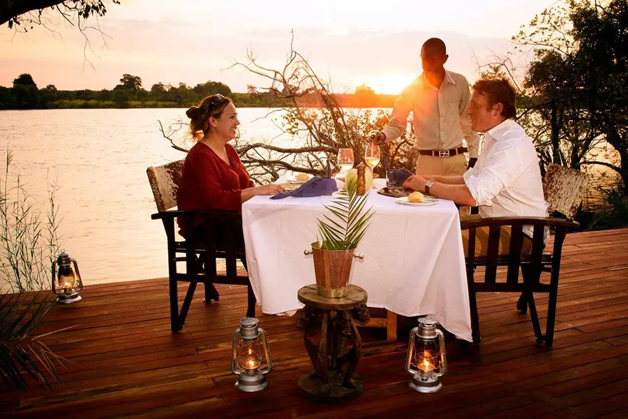 A couple sit at a table found on a deck by a river at sunset while another gentleman pours beverages for them | Go2Africa