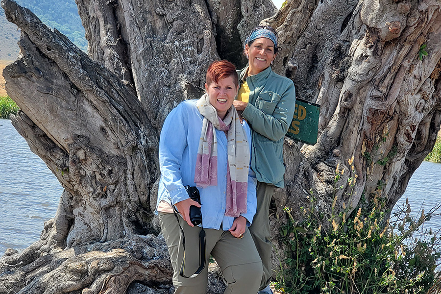A lesbian couple stands in front of an ancient tree while on safari in Tanzania, Africa. 