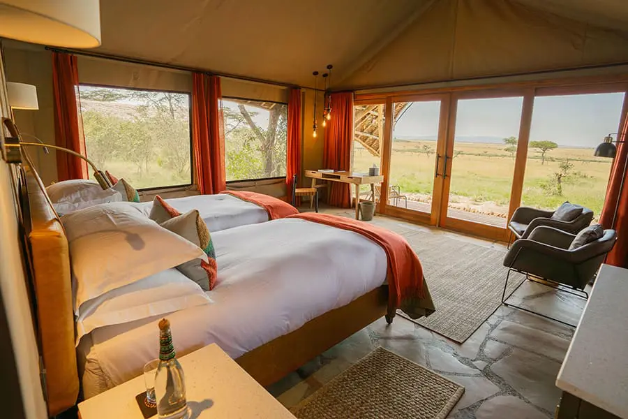 Twin beds in a spacious room with red decorative accents look out of expansive windows on two sides of the room and over the surrounding wilderness | Go2Africa