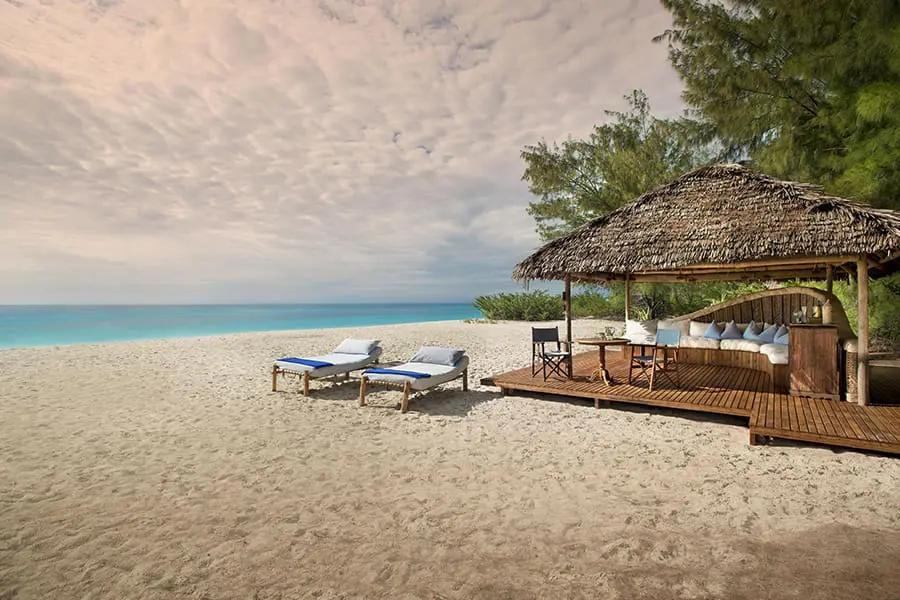 A thatched structure and platform below house a semicircular couch and small table and chairs while two sun loungers sit just outside on the beach | Go2Africa