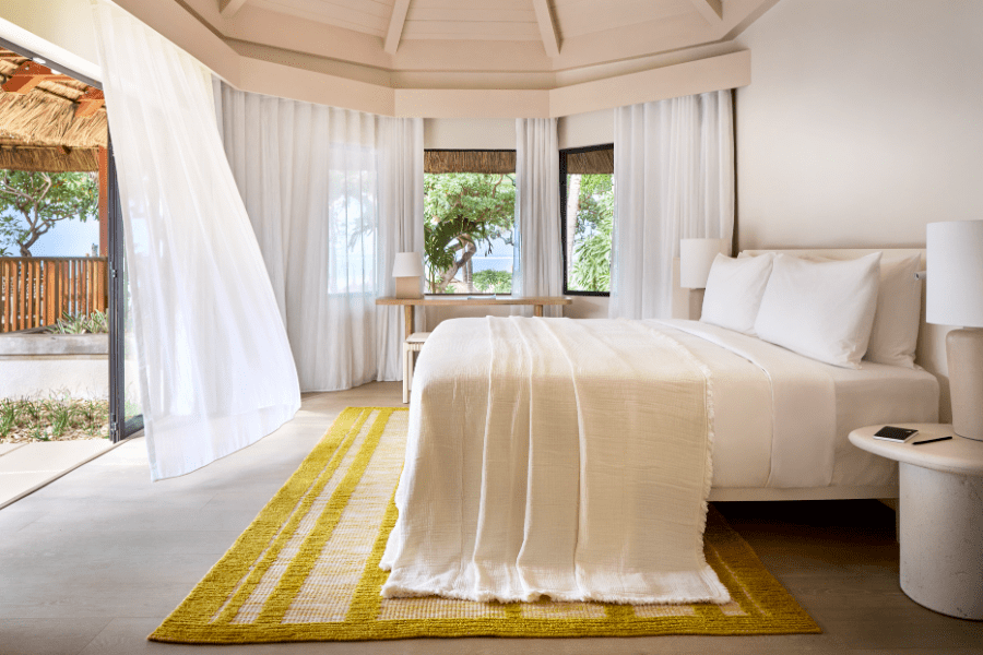 Suite with a double bed, yellow carpet and windows looking out to palm trees and a thatched roof at LUX* Belle Mare in Mauritius | Go2Africa