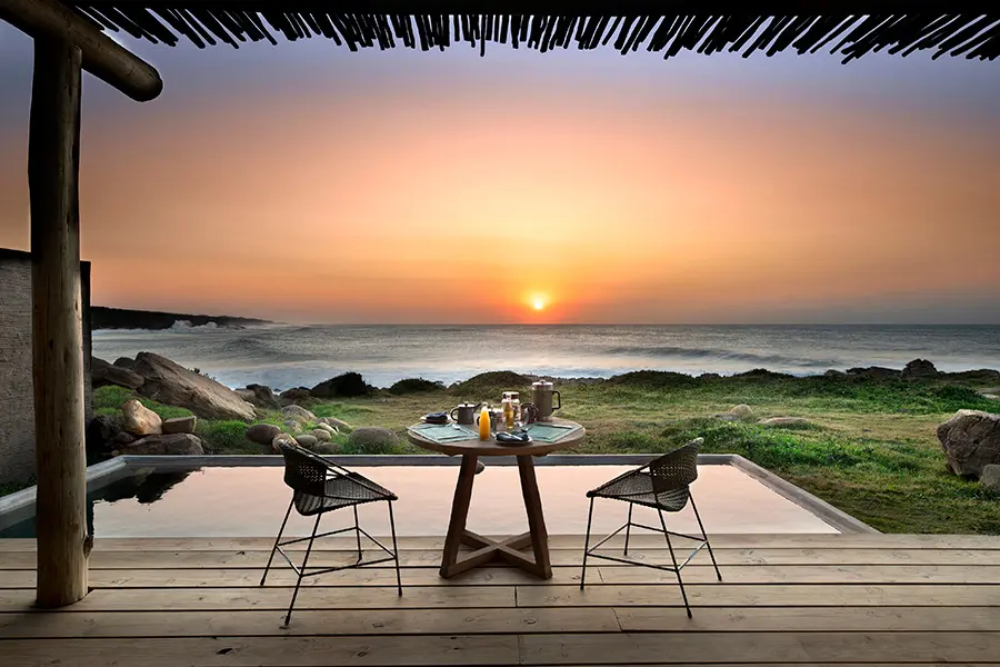 Dos sillas y una mesa en una terraza con vistas al océano y a la puesta de sol desde GweGwe Beach Lodge | Go2Africa