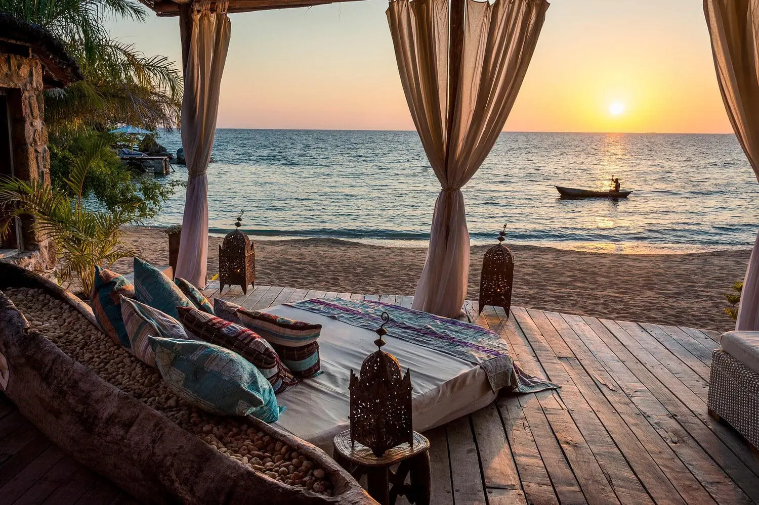 Una cama de día con vistas al lago Malawi al atardecer, donde a lo lejos, un velero flota suavemente en el agua.