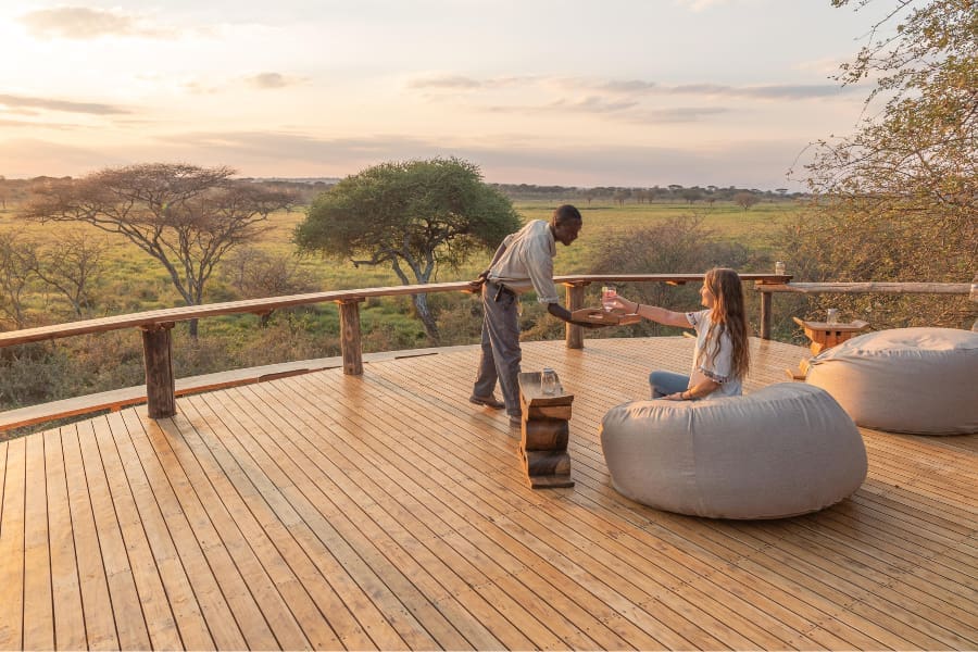 Una mujer acepta un trago al atardecer de uno de los miembros del equipo de Oliver's Camp en la terraza con una vista gloriosa de la puesta de sol | Go2Africa
